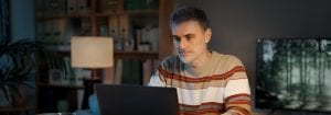 A man wearing a stripy jumper sits at his desk and is looking at the laptop in front of him